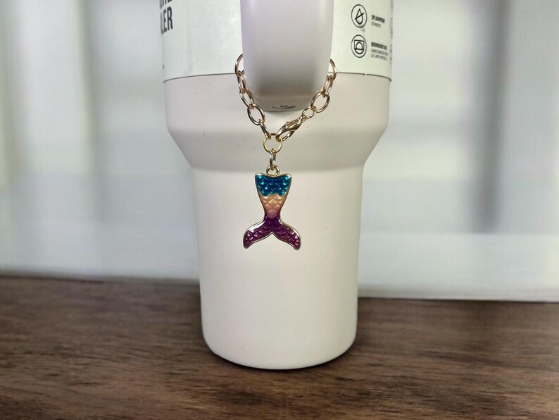 May include: A gold chain with a mermaid tail charm. The tail is made of metal and has a pink, blue, and yellow design.