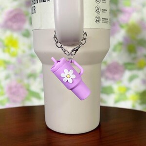 May include: A purple silicone tumbler charm with a white daisy attached to a silver chain. The charm is hanging from a stainless steel tumbler with the text "CHER H2.0 ER" on the side.