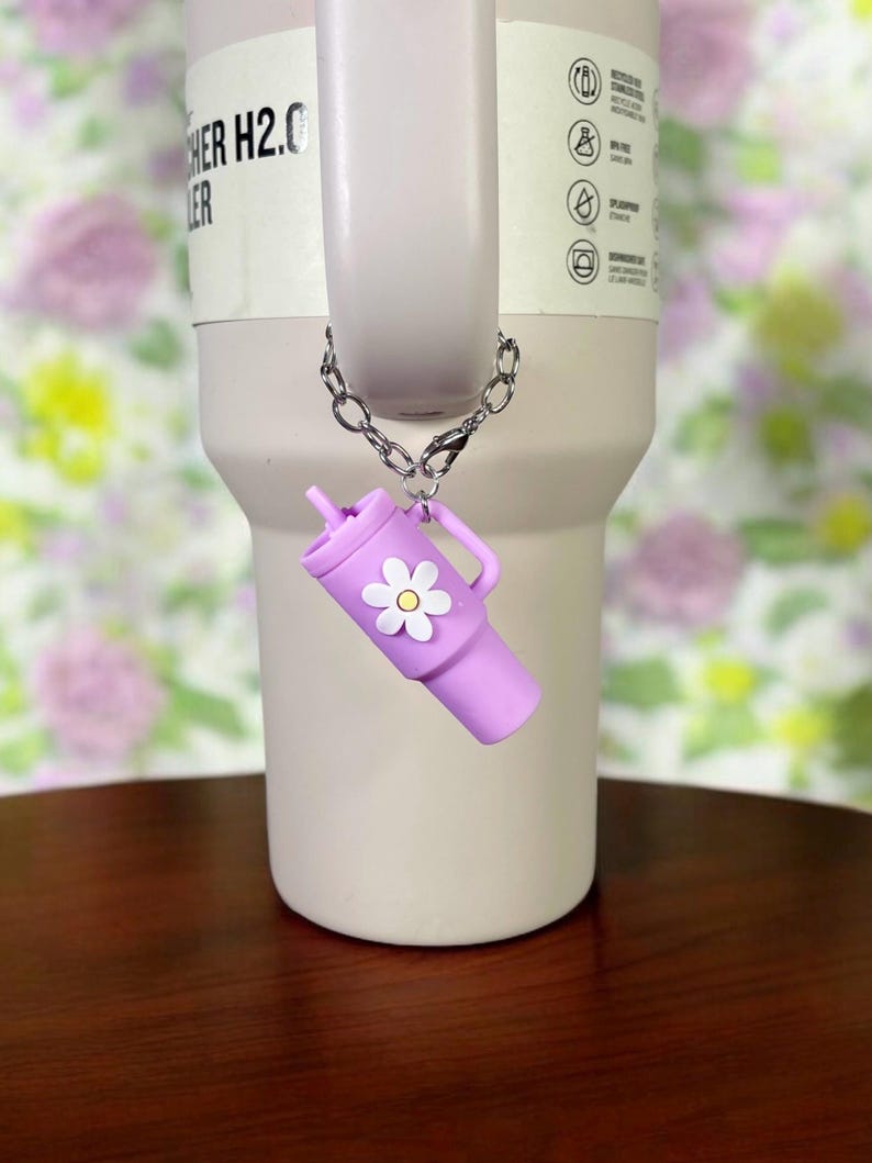 May include: A purple silicone tumbler charm with a white daisy attached to a silver chain. The charm is hanging from a stainless steel tumbler with the text "CHER H2.0 ER" on the side.