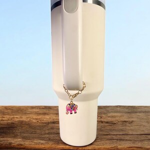May include: A white insulated tumbler with a handle, adorned with a gold chain and a pink elephant charm. The charm is detailed with colorful gems. The tumbler sits on a wooden surface against a light blue backdrop.