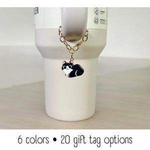 May include: A white travel mug with a black and white cat charm attached to a gold chain. The mug has the text "Cooler" on the side. The image text reads "6 colors • 20 gift tag options".