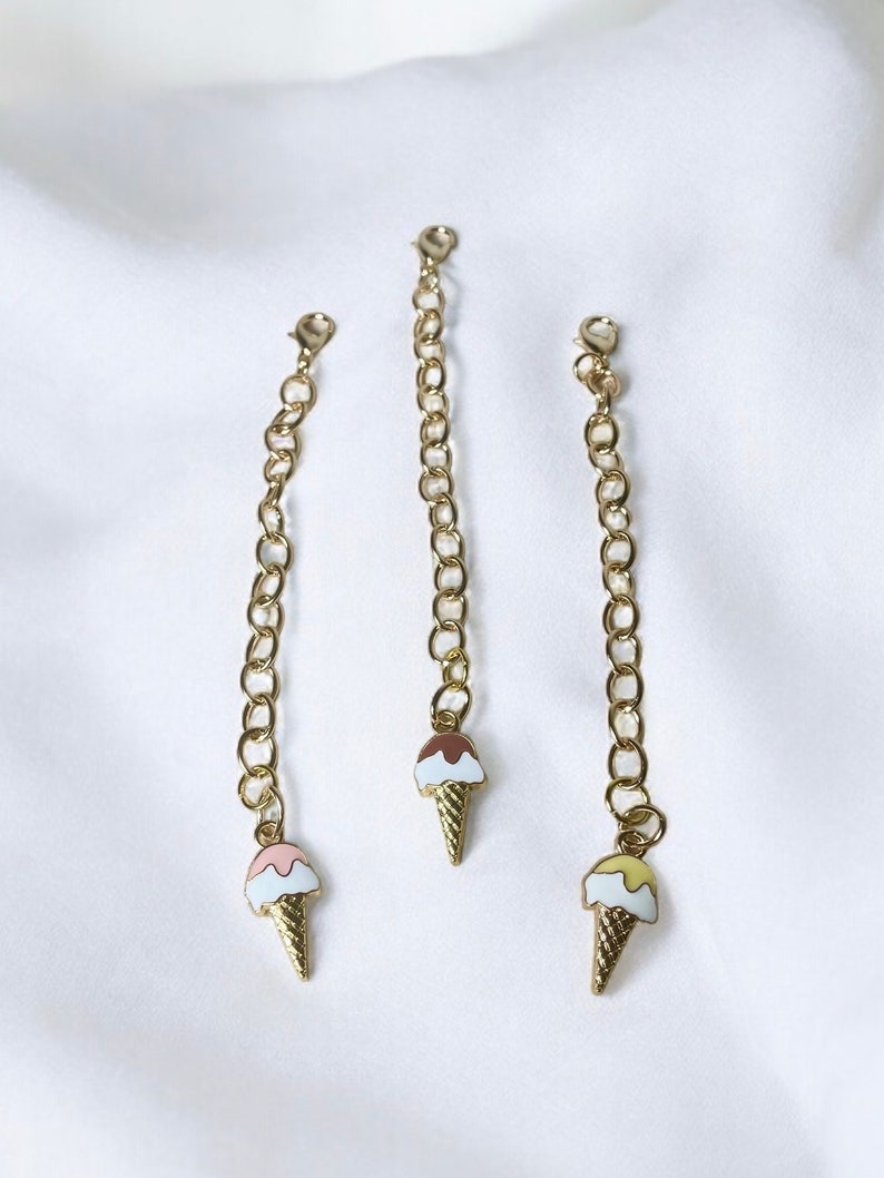 May include: Three gold chains with ice cream cone charms. The charms are different colours: pink, brown, and yellow.