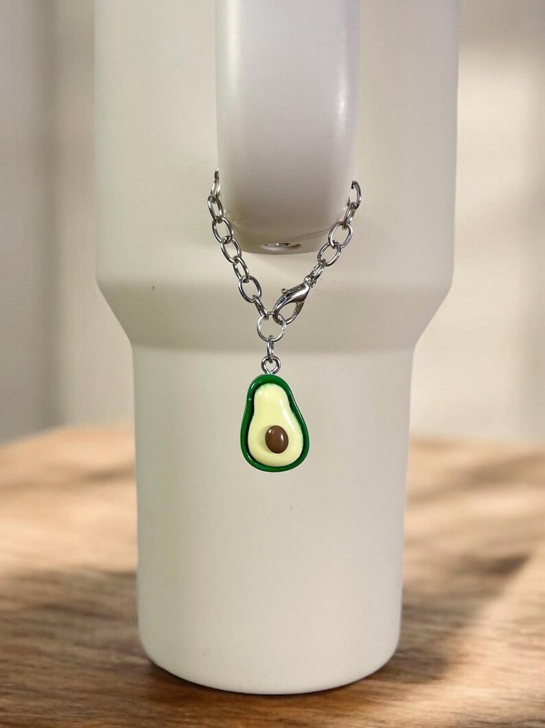 May include: A silver chain with a green and yellow avocado charm attached. The charm is hanging from a white tumbler.