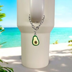 May include: A silver chain with a green avocado charm attached to a white tumbler. The tumbler is in front of a beach scene.