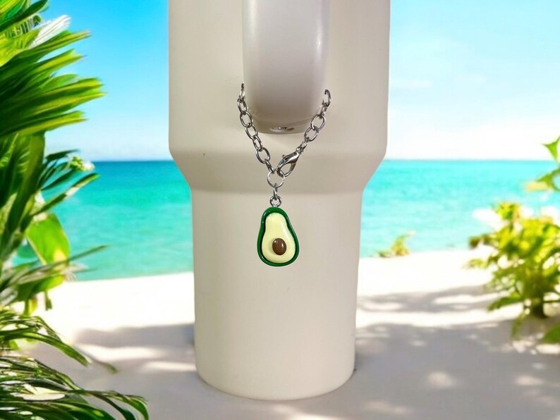 May include: A silver chain with a green avocado charm attached to a white tumbler. The tumbler is in front of a beach scene.