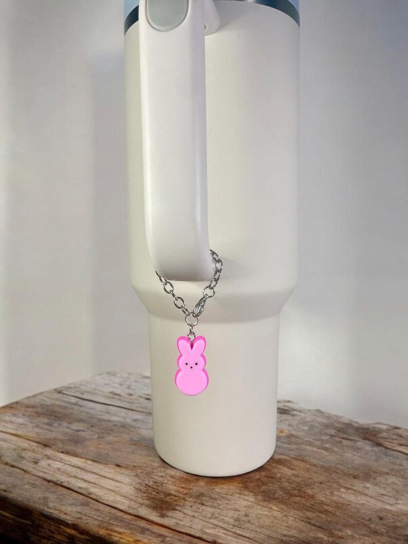 May include: A white travel mug with a pink bunny charm attached to the handle. The charm is on a silver chain.