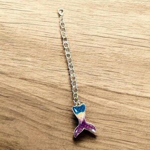 May include: A silver chain with a mermaid tail charm. The tail is made of resin and has a sparkly finish. The colors of the tail are blue, pink, and purple.