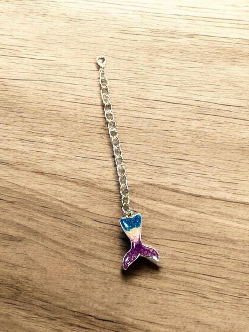 May include: A silver chain with a mermaid tail charm. The tail is made of resin and has a sparkly finish. The colors of the tail are blue, pink, and purple.