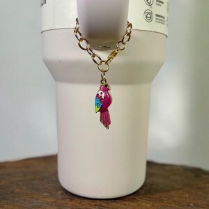 May include: A gold chain with a pink, blue, and green parrot charm attached. The charm is hanging from a white tumbler with the text "HER H2.0" on the side.