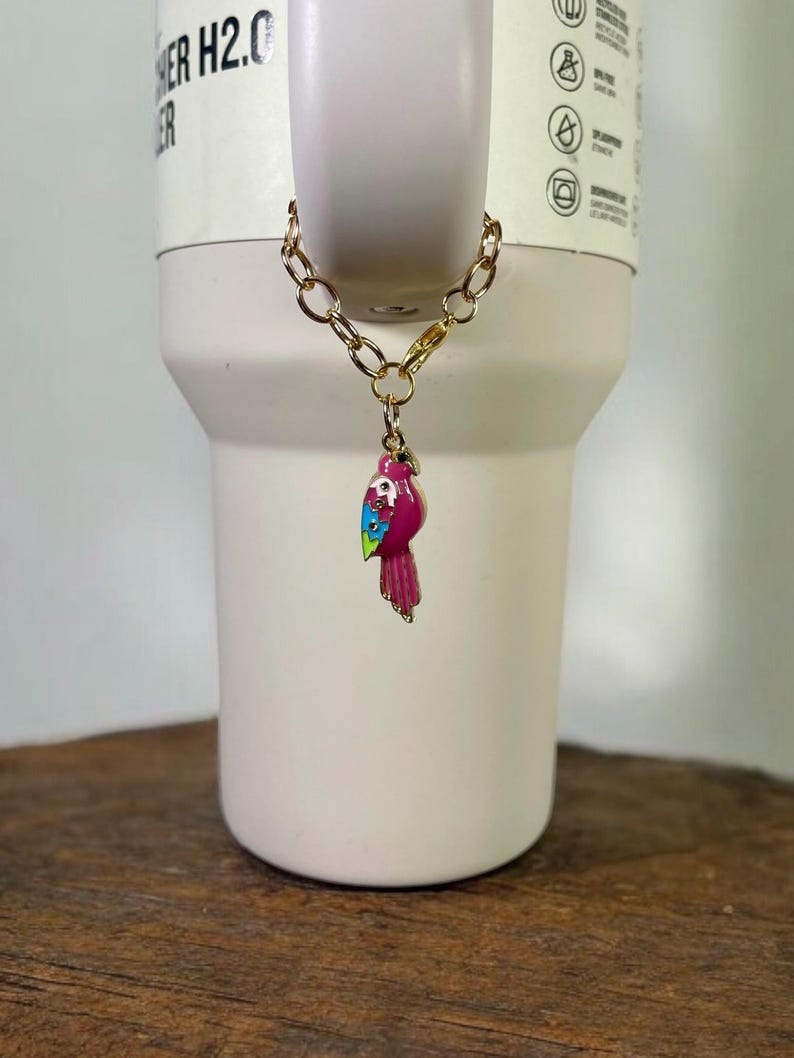 May include: A gold chain with a pink, blue, and green parrot charm attached. The charm is hanging from a white tumbler with the text "HER H2.0" on the side.