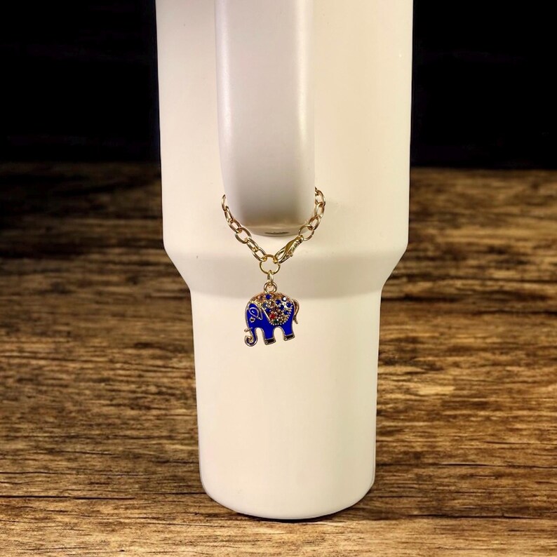 May include: A gold-colored chain with a blue and red elephant charm is attached to a white tumbler. The elephant charm has a blue enamel body with red and clear crystal accents. The tumbler is white and has a handle.