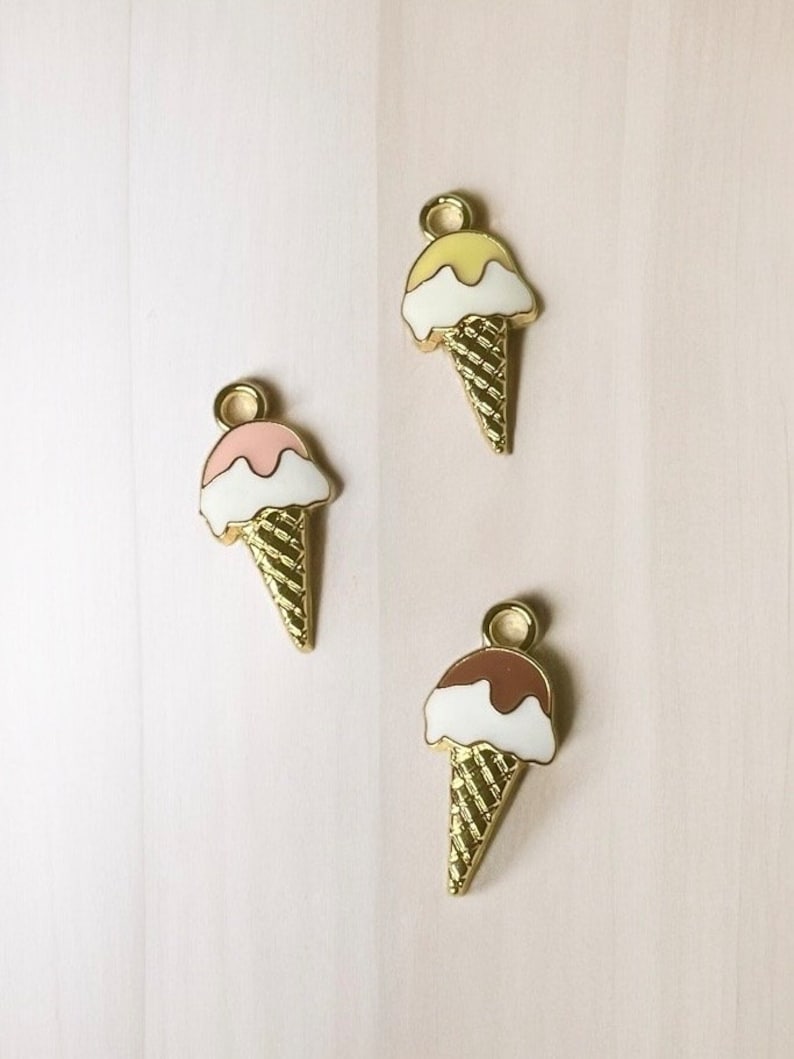 May include: Three gold-coloured ice cream cone charms with different coloured ice cream scoops. The ice cream scoops are yellow, pink, and brown.