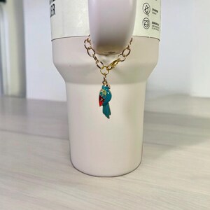 May include: A gold chain with a blue and red bird charm attached to a white travel mug.