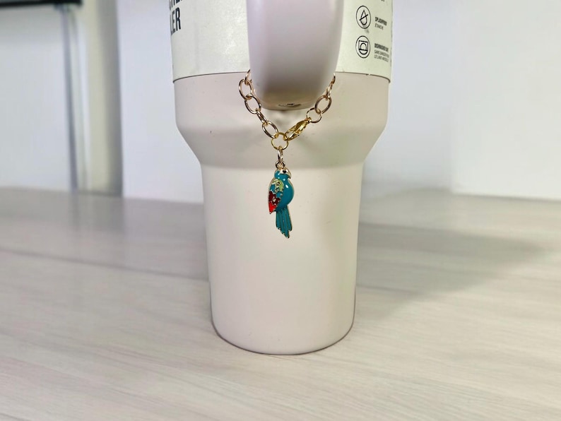 May include: A gold chain with a blue and red bird charm attached to a white travel mug.