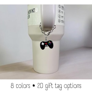Könnte beinhalten: Eine weiße Reise-Thermoskanne mit einem schwarzen Videospiel-Controller-Anhänger, der mit einer silbernen Kette befestigt ist. Die Tasse hat den Text "THER H2O" an der Seite. 8 Farben und 20 Geschenk-Tag-Optionen sind verfügbar.