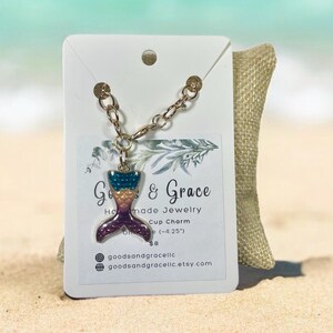 May include: A gold chain with a mermaid tail charm. The charm is purple, teal, and yellow with glitter accents. The charm is 4.25 inches long. The chain is 8 dollars.
