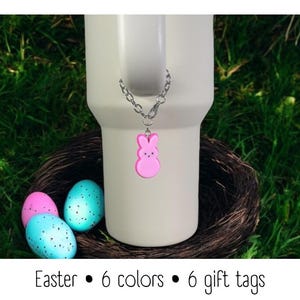May include: Pink bunny shaped charm on a silver chain attached to a white travel mug. The mug is sitting in a bird's nest with blue and pink Easter eggs.