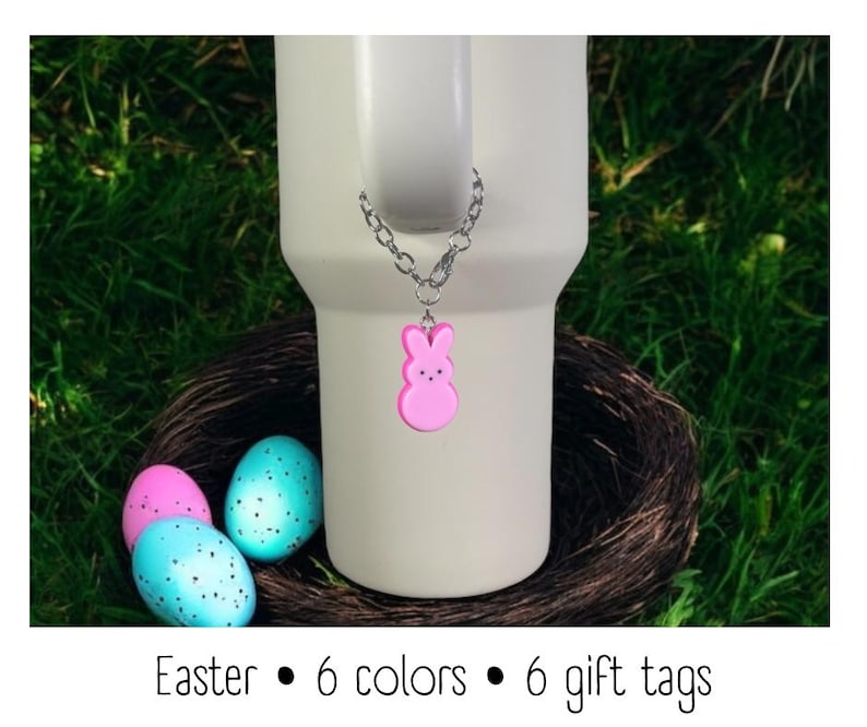 May include: Pink bunny shaped charm on a silver chain attached to a white travel mug. The mug is sitting in a bird's nest with blue and pink Easter eggs.