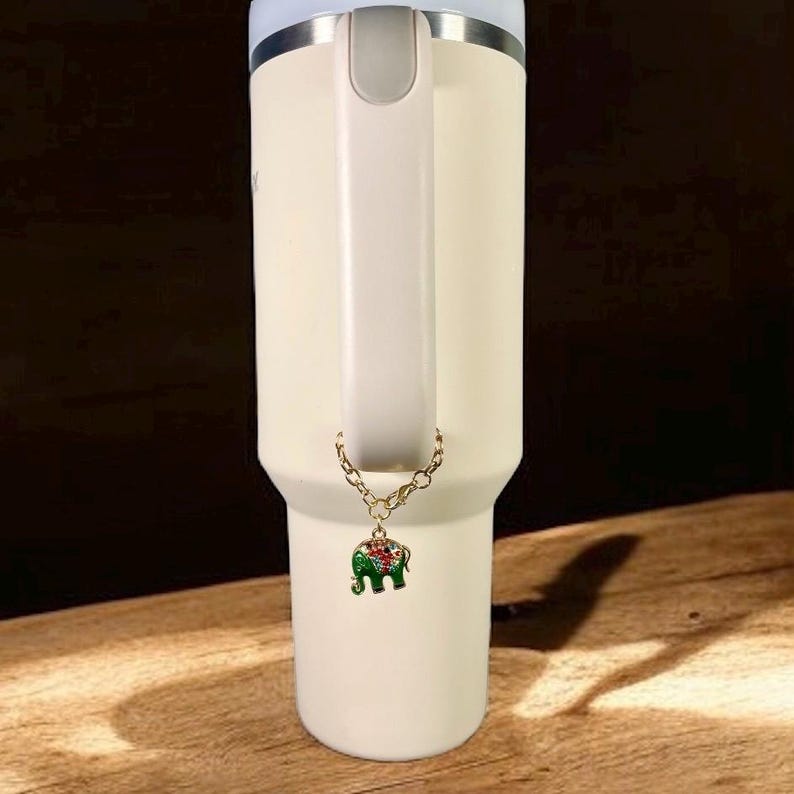 May include: A cream-colored insulated tumbler with a handle and a gold-colored chain charm. The charm is an enameled green elephant with red and white accents. The tumbler has a silver rim and a white handle.
