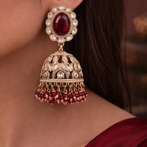 May include: Elaborate gold-coloured earrings featuring a large, oval, ruby-red gemstone surrounded by smaller white stones. The lower section is bell-shaped, decorated with white stones and dangling red beads. A stunning piece of jewellery.