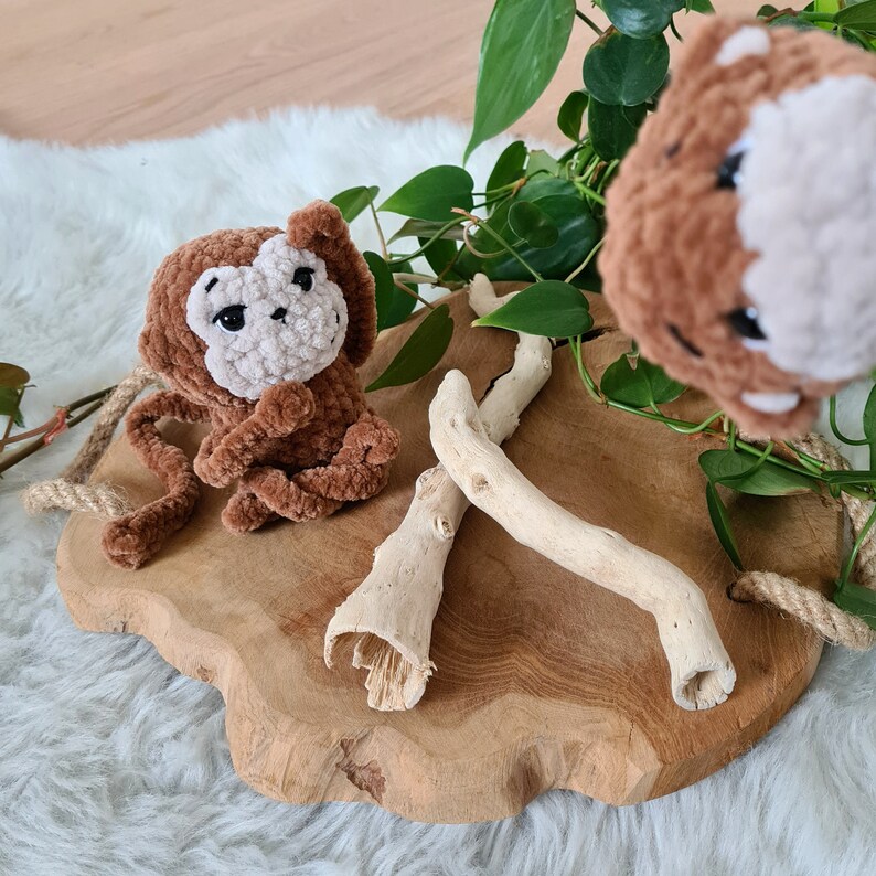May include: Two brown crocheted monkey toys on a wooden surface with a green plant and driftwood.