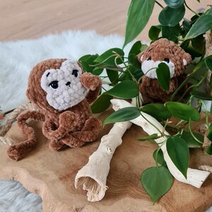 May include: Two brown crocheted monkey toys sitting on a wooden surface with green leafy plants in the background.
