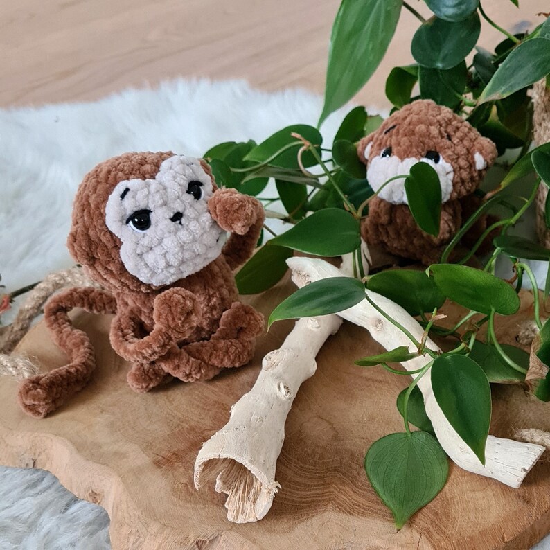 May include: Two brown crocheted monkey toys sitting on a wooden surface with green leafy plants in the background.