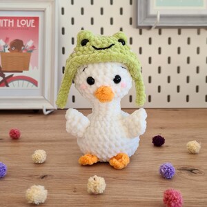 Pode incluir: Pato branco de croch&ecirc; usando um chap&eacute;u de sapo verde. O pato est&aacute; sentado em uma superf&iacute;cie de madeira com pompons coloridos espalhados ao redor.