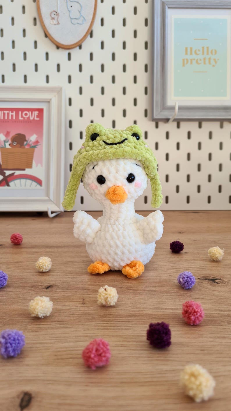 Pode incluir: Pato branco de croch&ecirc; usando um chap&eacute;u de sapo verde. O pato est&aacute; sentado em uma superf&iacute;cie de madeira com pompons coloridos espalhados ao redor.