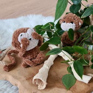 May include: Two brown crocheted monkey toys. One monkey is covering its eyes with its hands. The monkeys are sitting on a wooden surface with a white branch and green leaves.