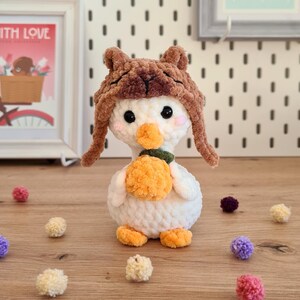 Pode incluir: Um pato branco de croch&ecirc; usando um chap&eacute;u de croch&ecirc; marrom com abas de orelha felpudas. O pato est&aacute; segurando uma ab&oacute;bora de croch&ecirc; laranja. O pato est&aacute; cercado por pompons coloridos.