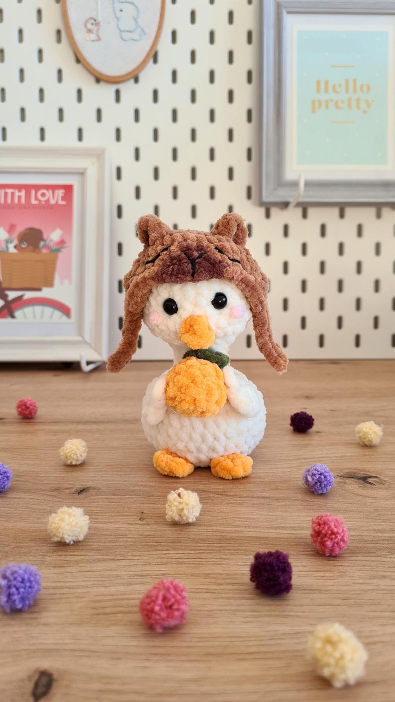 Pode incluir: Um pato branco de croch&ecirc; usando um chap&eacute;u de croch&ecirc; marrom com abas de orelha felpudas. O pato est&aacute; segurando uma ab&oacute;bora de croch&ecirc; laranja. O pato est&aacute; cercado por pompons coloridos.