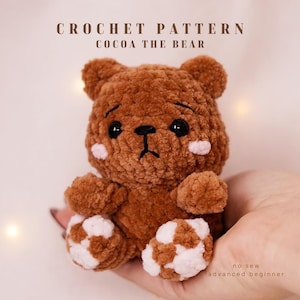 Könnte beinhalten: Ein handgefertigter brauner Häkelbär mit weiß-braun karierten Pfoten und rosa Wangen. Der Bär hat schwarze Augen und eine schwarz gestickte Nase und Mund. Der Text oben lautet "CROCHET PATTERN COCOA THE BEAR". Der Text unten lautet "no sew advanced beginner".