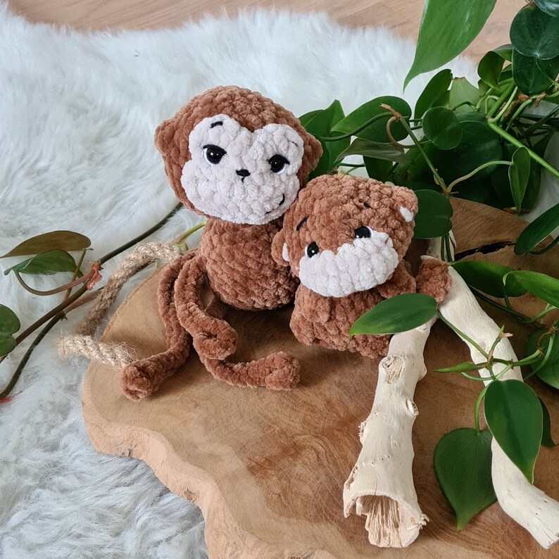 May include: Two brown crocheted monkey toys, one larger than the other, sitting on a wooden surface. The smaller monkey is sitting on top of the larger monkey's back.