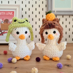 Pode incluir: Dois peluches de pato branco de croch&ecirc; usando chap&eacute;us. Um pato est&aacute; usando um chap&eacute;u de sapo verde e o outro est&aacute; usando um chap&eacute;u marrom peludo com uma pompom laranja no topo. Os patos est&atilde;o sentados em uma superf&iacute;cie de madeira com pompons coloridos espalhados ao redor.
