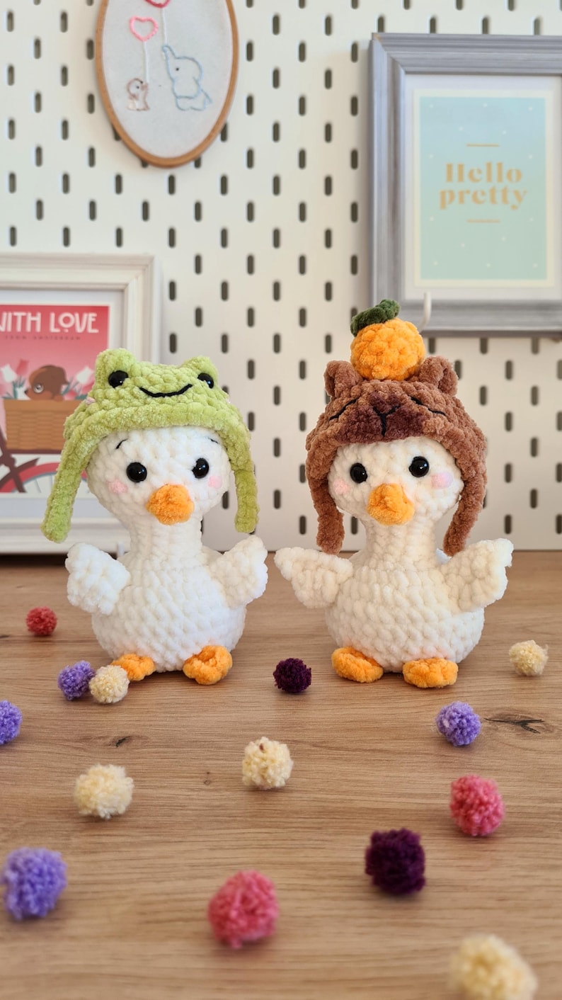 Pode incluir: Dois peluches de pato branco de croch&ecirc; usando chap&eacute;us. Um pato est&aacute; usando um chap&eacute;u de sapo verde e o outro est&aacute; usando um chap&eacute;u marrom peludo com uma pompom laranja no topo. Os patos est&atilde;o sentados em uma superf&iacute;cie de madeira com pompons coloridos espalhados ao redor.