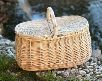 May include: A natural woven wicker picnic basket with a hinged lid and a carrying handle. The basket is oval shaped and has a light brown color.