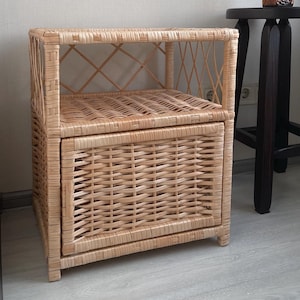 Puede incluir: Una mesa auxiliar de mimbre de color marrón claro con un cajón y un estante abierto. La mesa tiene un diseño tejido y está sobre un suelo de madera clara.