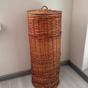 Puede incluir: Un cesto cilíndrico alto de mimbre con un acabado marrón natural. La cesta presenta un patrón trenzado en todo su cuerpo e incluye una tapa circular a juego con una pequeña asa trenzada en la parte superior para facilitar su apertura.