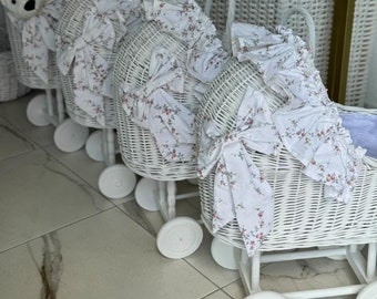 Puede incluir: Cuatro cochecitos de muñecas de mimbre blanco con ribetes de encaje blanco y tela con estampado floral. Los cochecitos tienen ruedas blancas y están alineados en fila.