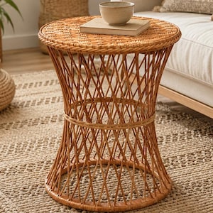 May include: A round side table constructed from woven light brown rattan, featuring a flared hourglass-shaped base with a crisscross lattice pattern and a flat circular top surface, supporting a small beige ceramic bowl resting on a book.