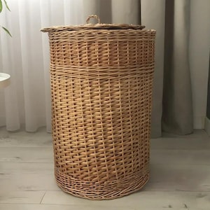 May include: A tall, cylindrical wicker laundry hamper with a lid. The hamper is made of woven light brown wicker and has a rounded base. A small, round handle is on the lid. The hamper is next to a white table with a potted plant.