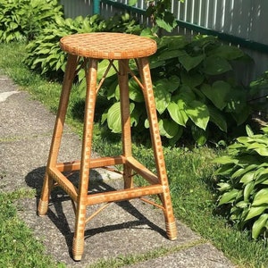 May include: A woven wicker stool with a square base and a round seat. The stool is brown and has a natural finish. It is sitting on a concrete patio surrounded by green grass and plants.