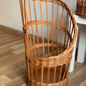 May include: A tall, cylindrical wicker basket featuring a spiralling open-frame design. The structure is crafted from light brown woven willow branches, with a solid circular base and vertical slats that curve upwards to form a high, slanted rim.