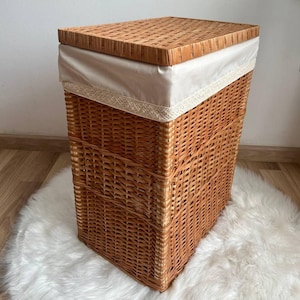 Puede incluir: Una cesta de lavandería rectangular de mimbre con tapa y forro de tela blanco. La tapa tiene un patrón tejido y el forro tiene un borde de encaje.