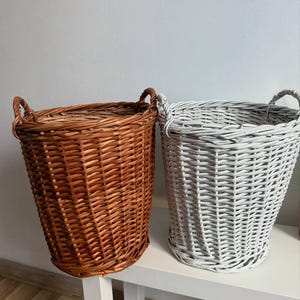 Pode incluir: Duas cestas de vime tecidas. Uma é de cor castanha quente e a outra é pintada de branco. Ambas têm alças arredondadas e uma forma cónica. As cestas são provavelmente usadas para armazenamento ou decoração.