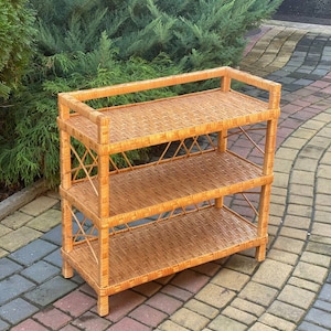 May include: A three-tiered wicker shelf unit with a natural brown finish. The shelf is made of woven wicker and has a simple, rustic design.