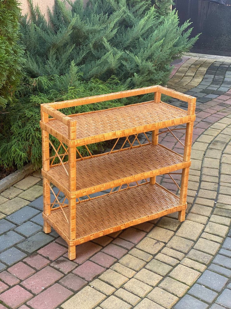 May include: A three-tiered wicker shelf unit with a natural brown finish. The shelf is made of woven wicker and has a simple, rustic design.