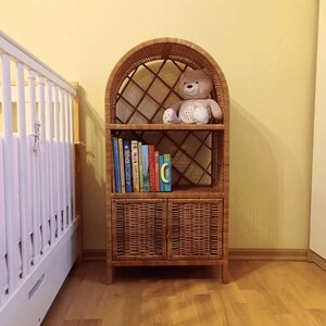 Puede incluir: Una estantería de mimbre con parte superior arqueada y dos puertas. La estantería está llena de libros y un peluche.