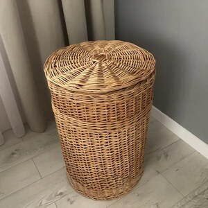 May include: A tall, cylindrical wicker laundry hamper with a domed lid. The hamper is made of woven light brown wicker and sits on a light-colored wooden floor. The lid has a central knob.
